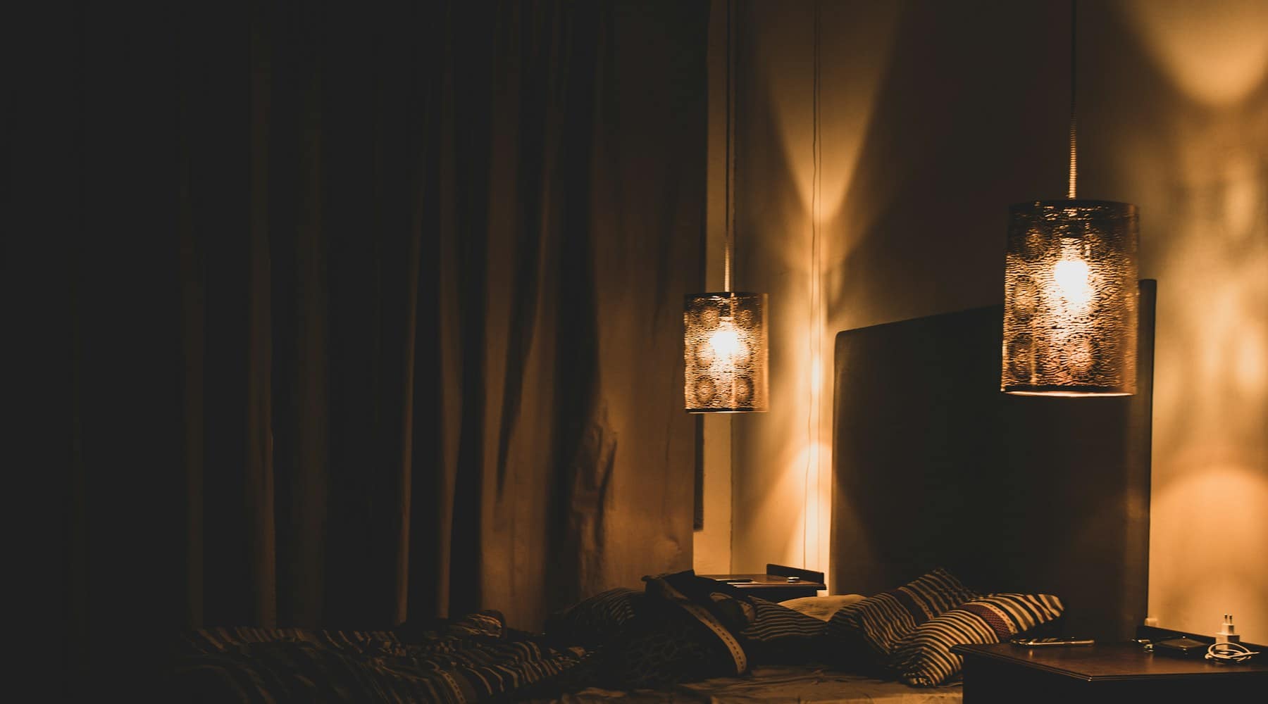 A dark bedroom lit only by two amber pendant lamps beside an unmade bed